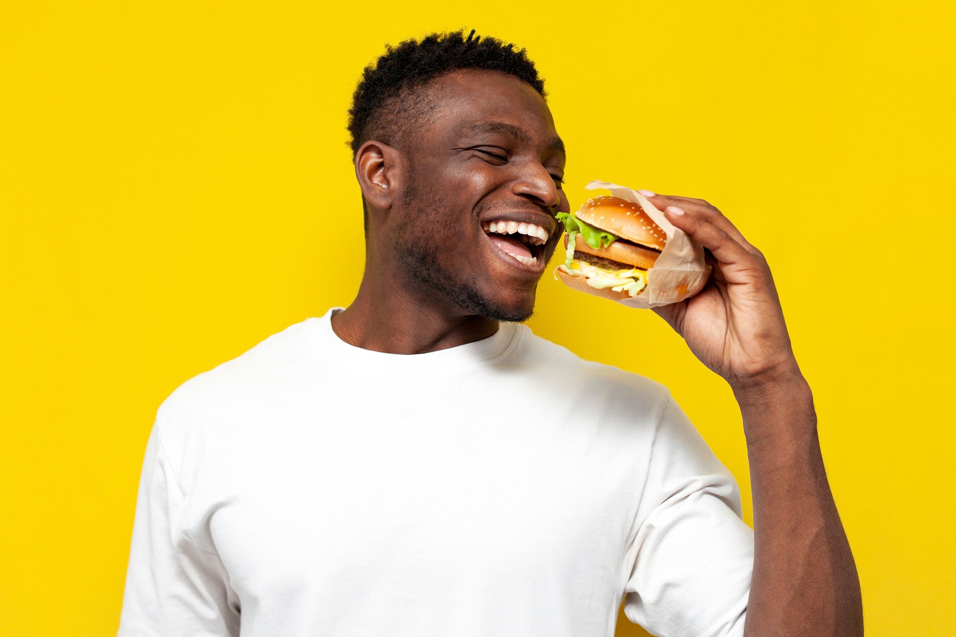 african american man in white t-shirt holding big burger and smiling, the guy eats fast food on yellow background